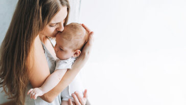 Woman holding a baby and kissing the baby on the head in front of a white background.