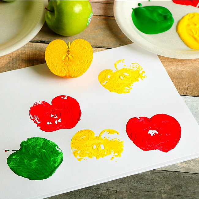 Red, green, and yellow apple shapes stamped on a white canvas sitting on a wood table with two apples and two paper plates in honor of Fall.