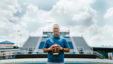 Coach Jeff Conaway holding a football, standing in the middle of the TFA football field.