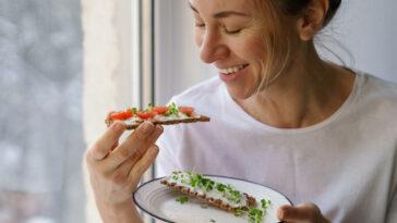 Smiling woman eating rye crisp bread with creamy vegetarian cheese tofu, cherry tomato and rucola micro greens, sitting at home and looking at window. Healthy food, gluten free, diet concept.